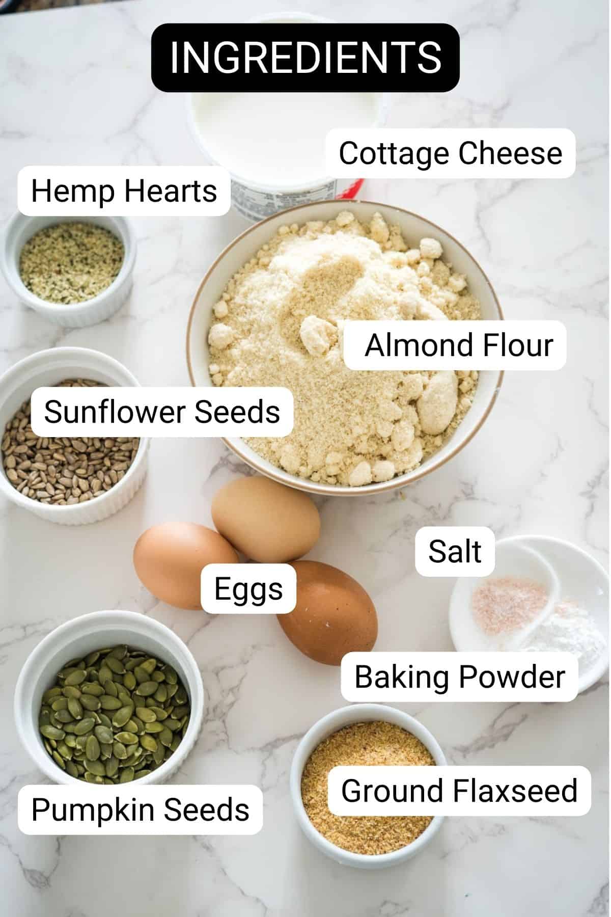 A marble countertop with ingredients labeled: cottage cheese, almond flour, sunflower seeds, hemp hearts, pumpkin seeds, ground flaxseed, salt, baking powder, and eggs.