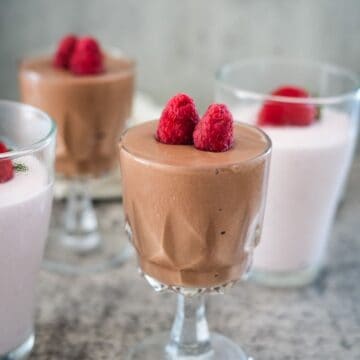 Four dessert glasses with mousse, two with chocolate mousse topped with raspberries and two with pink mousse also topped with raspberries. They are placed on a gray textured surface.