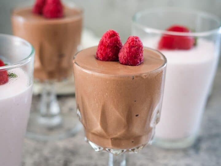 Four dessert glasses with mousse, two with chocolate mousse topped with raspberries and two with pink mousse also topped with raspberries. They are placed on a gray textured surface.