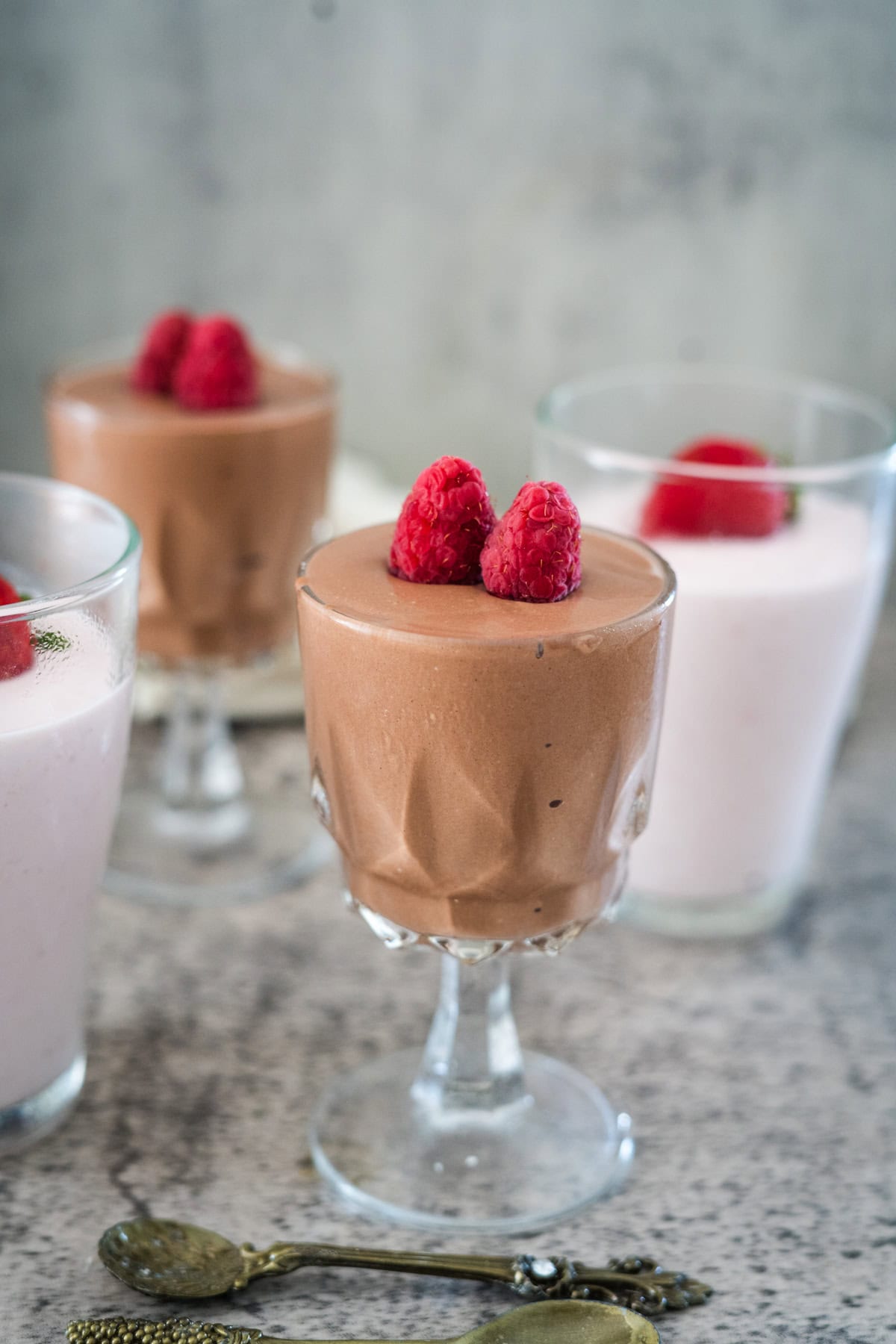 Four glass cups filled with chocolate and strawberry mousse topped with raspberries, arranged on a grey surface with two small spoons nearby.