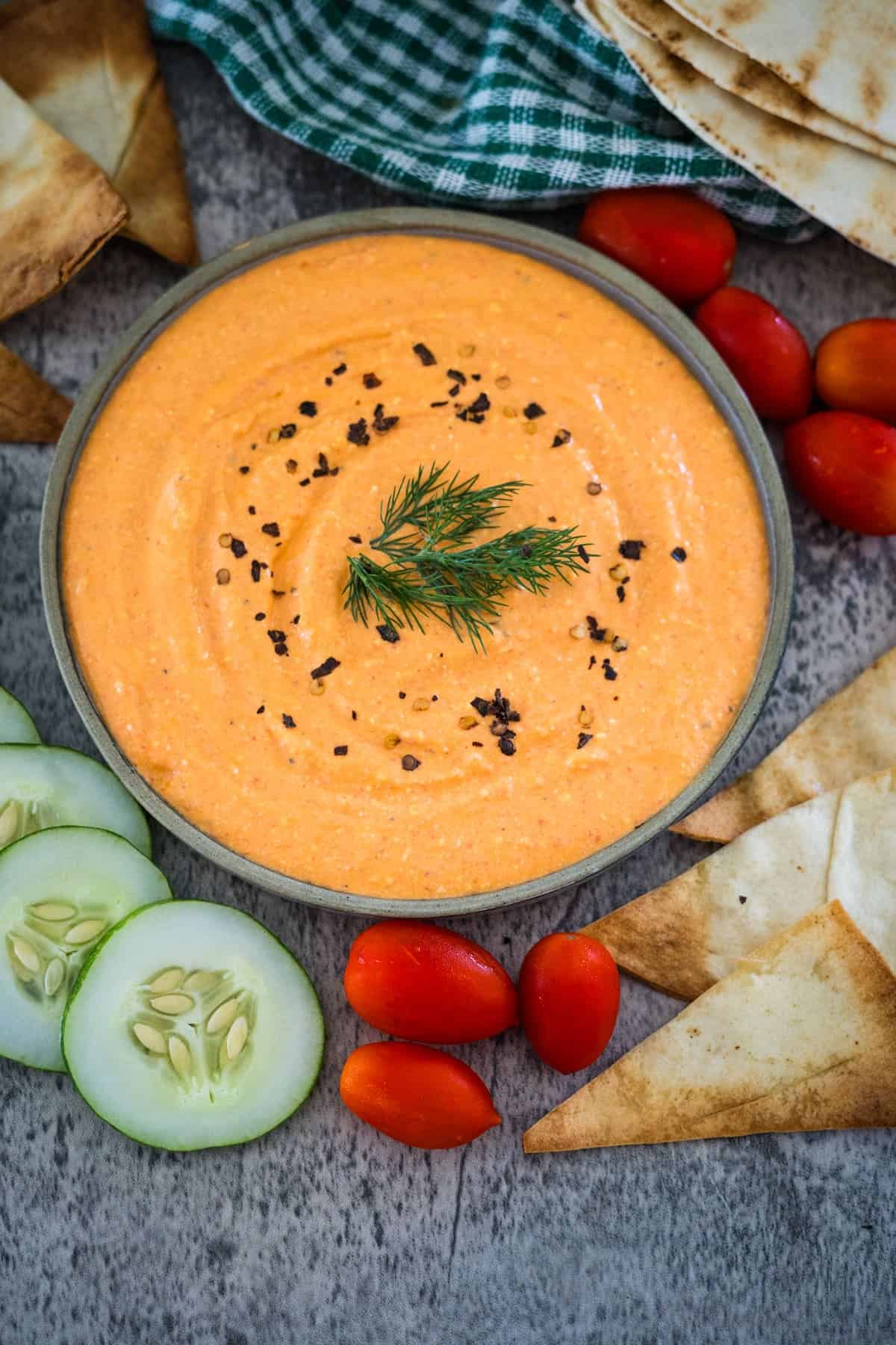 A bowl of creamy orange dip garnished with dill and black pepper flakes, surrounded by pita chips, cucumber slices, and cherry tomatoes on a gray surface with a green checkered cloth.