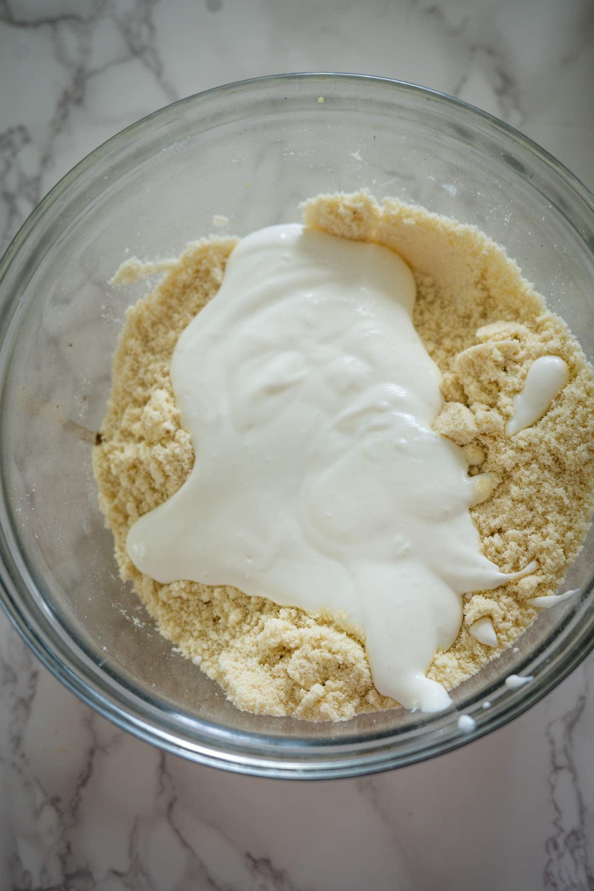 A glass bowl containing a mixture of flour and a white liquid, perhaps prepping for some delicious cottage cheese cookies, being combined on a marble countertop.