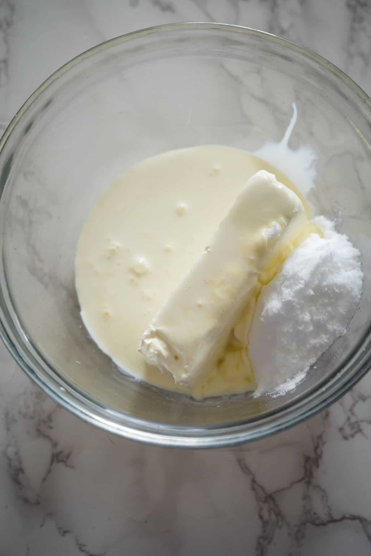 A clear glass bowl containing a stick of butter, a pile of powdered sugar, and a liquid ingredient, likely milk or cream, sits on a marble countertop—a scene reminiscent of preparing delightful nutella brownie cheesecake.