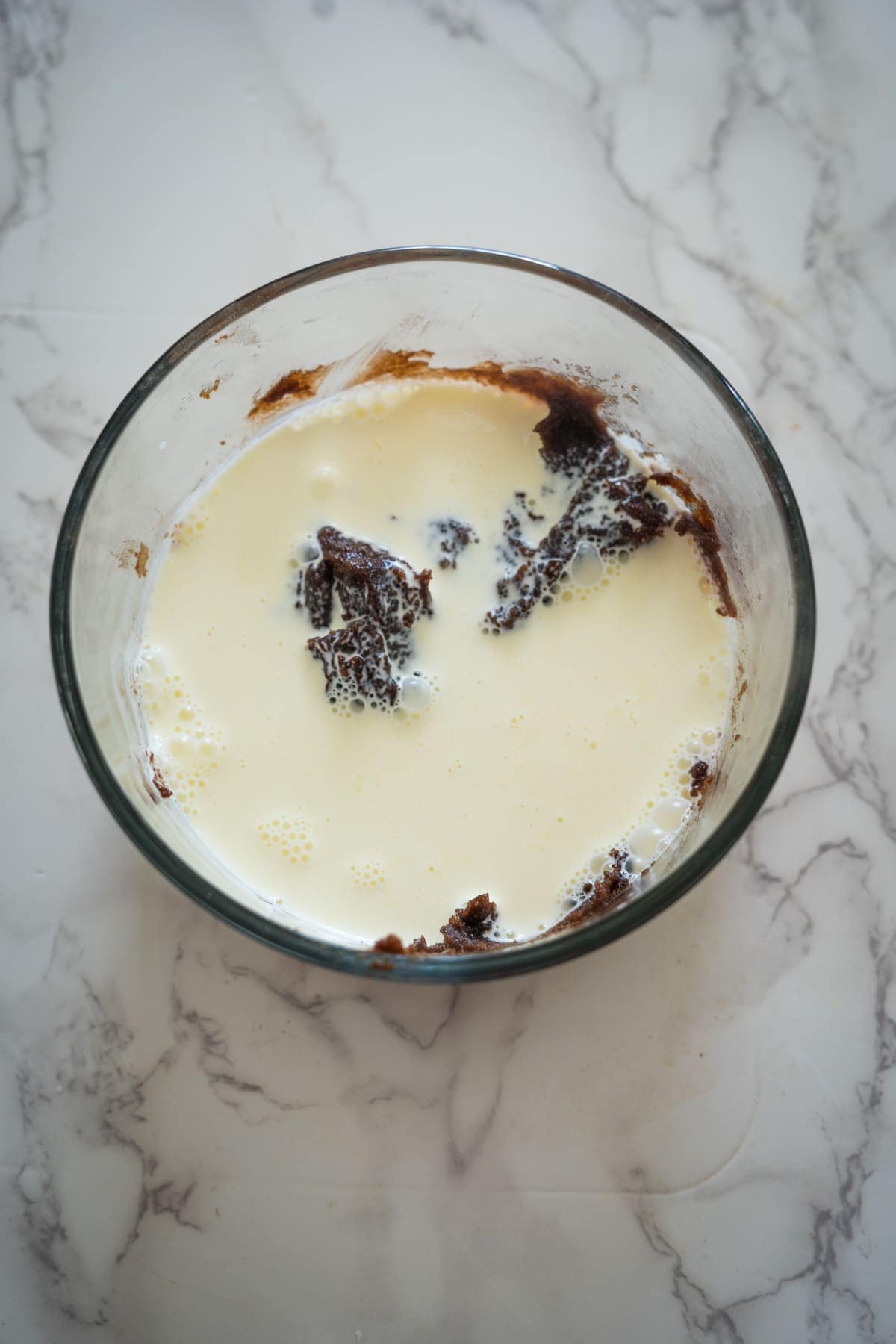 A clear glass bowl containing a mixture of cream and dark, crumbly Nutella brownie pieces rests on a marble countertop.