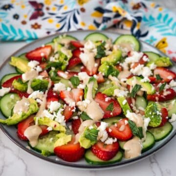 A fresh salad with sliced strawberries, cucumbers, and crumbled cheese, garnished with herbs and drizzled with a creamy dressing, served on a plate with a floral napkin alongside.