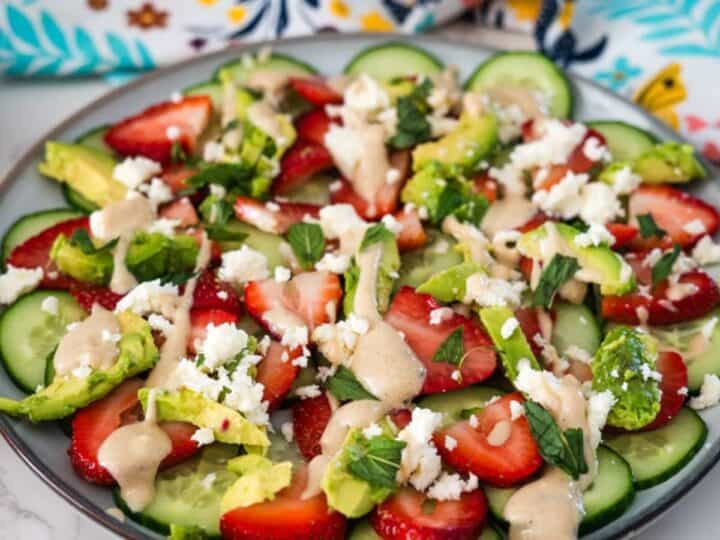 A fresh salad with sliced strawberries, cucumbers, and crumbled cheese, garnished with herbs and drizzled with a creamy dressing, served on a plate with a floral napkin alongside.