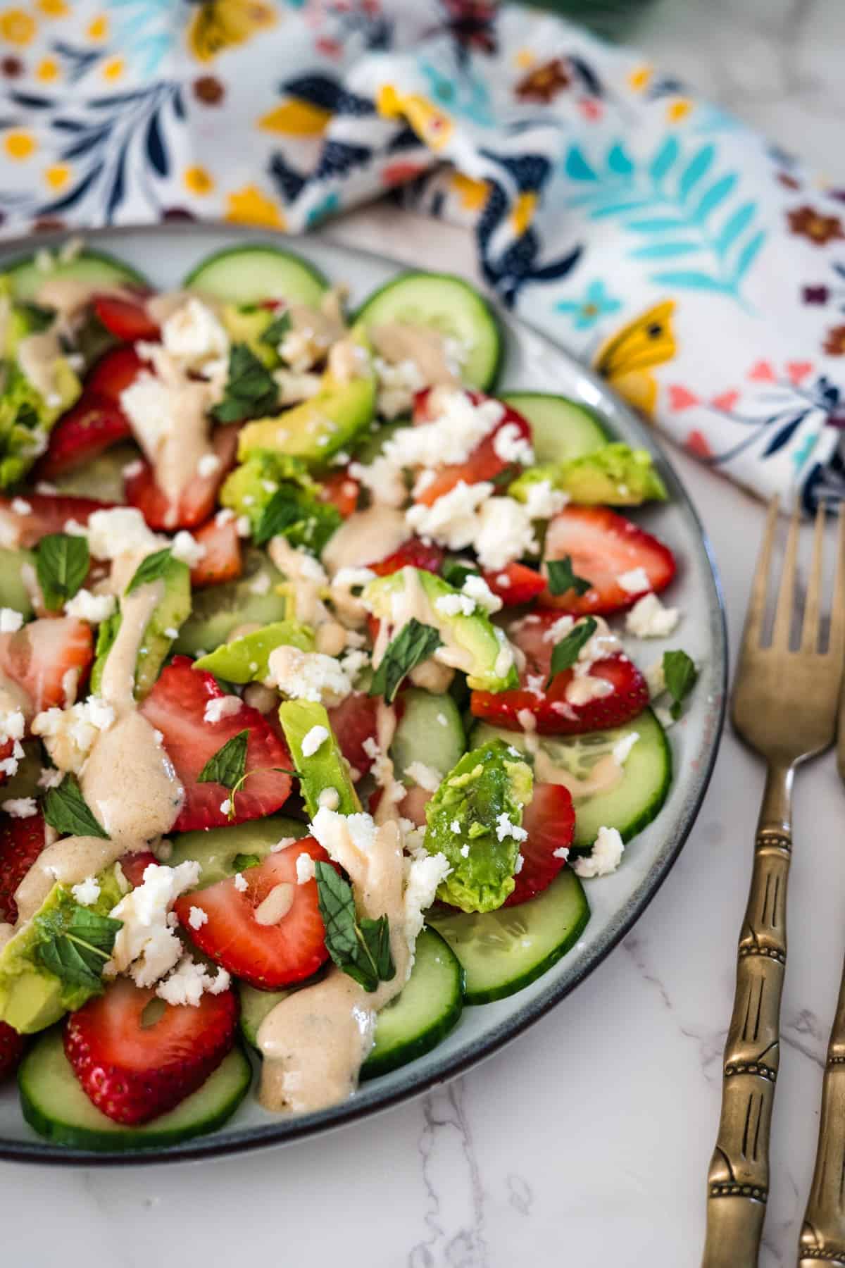 A vibrant salad with sliced strawberries, cucumbers, avocado, and crumbled cheese on a decorative plate beside ornate cutlery.