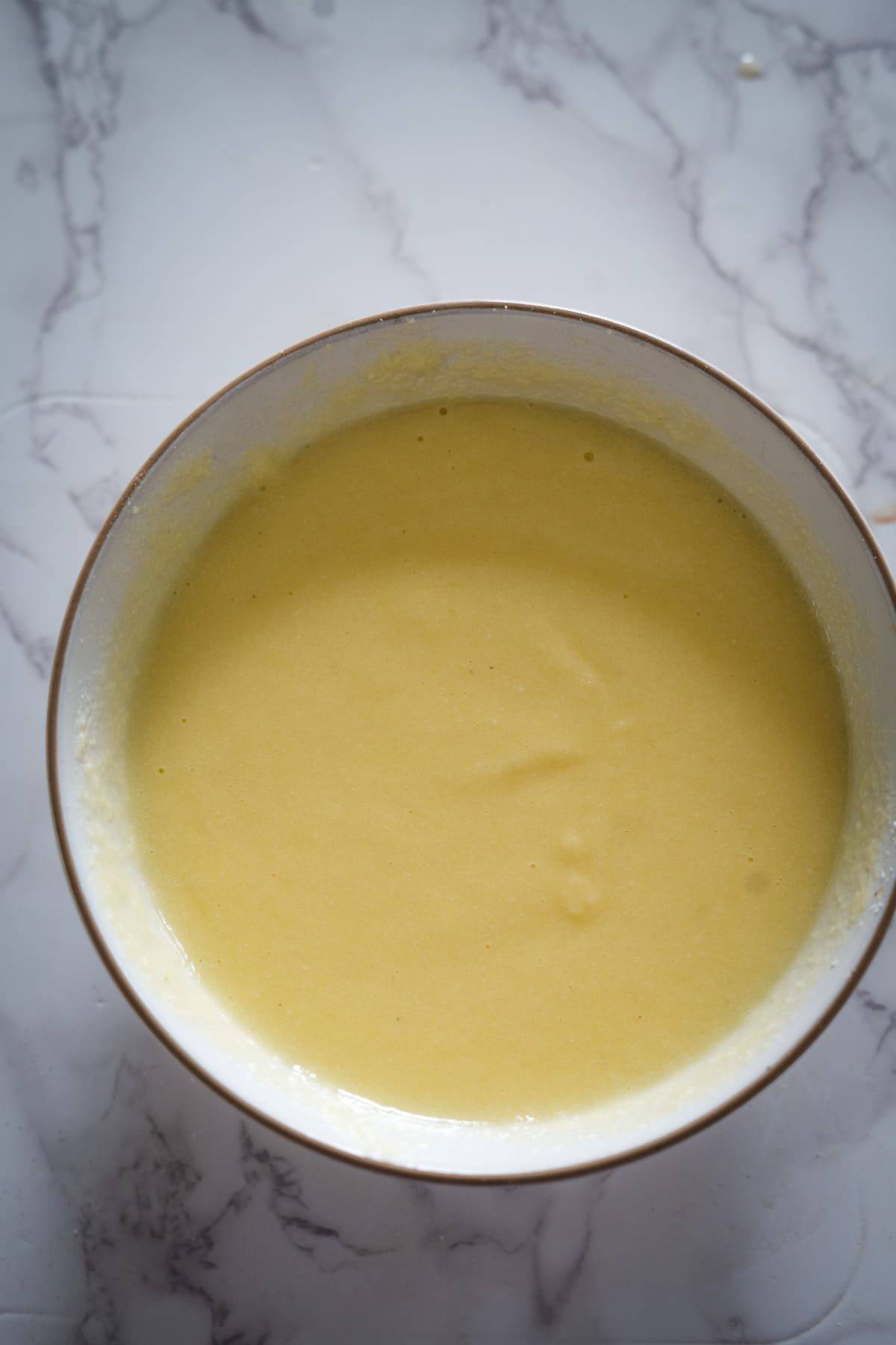 A bowl filled with a smooth, light yellow batter, perfect for lemon lavender cupcakes, is placed on a white marble surface.