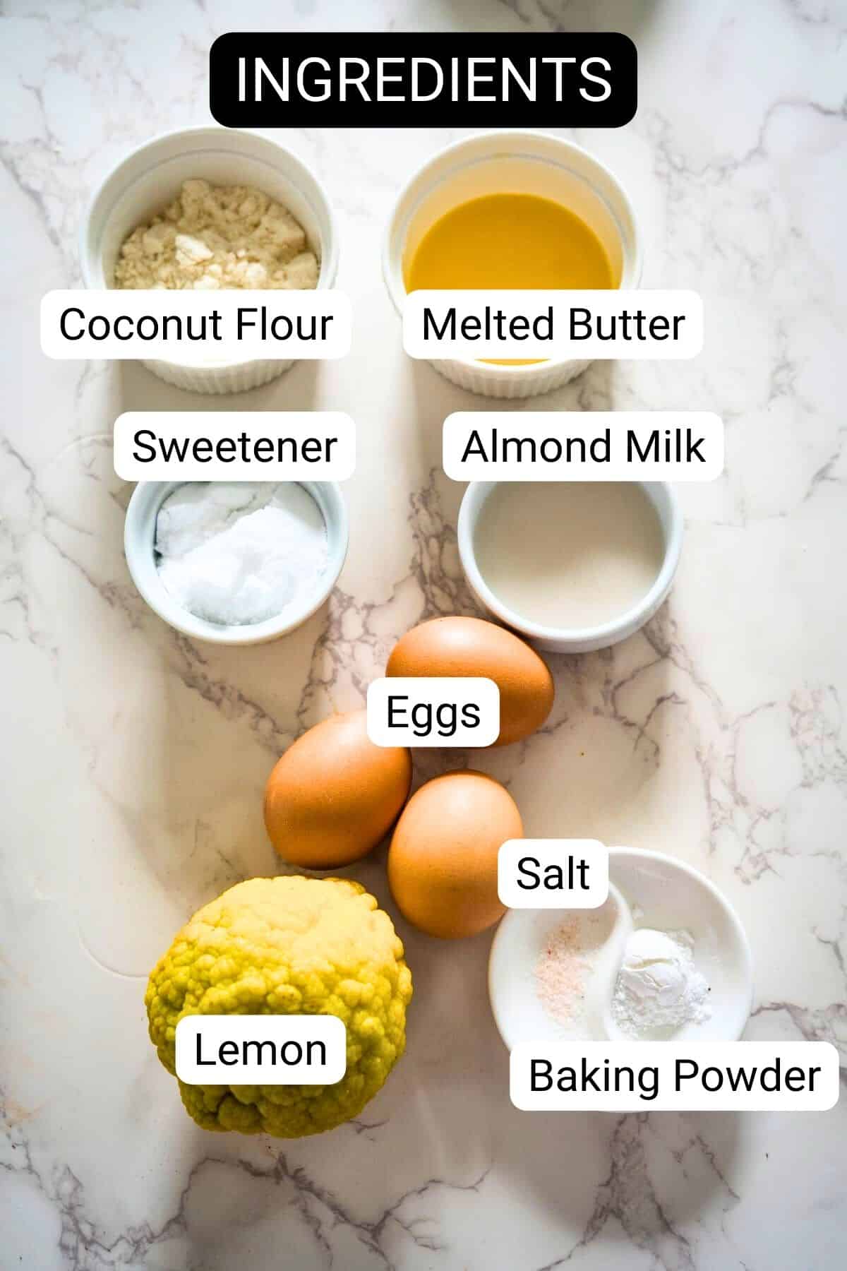 Ingredients arranged on a marble countertop include coconut flour, sweetener, almond milk, melted butter, eggs, salt, baking powder, and a lemon. Each item is labeled for making delicious lemon lavender cupcakes.