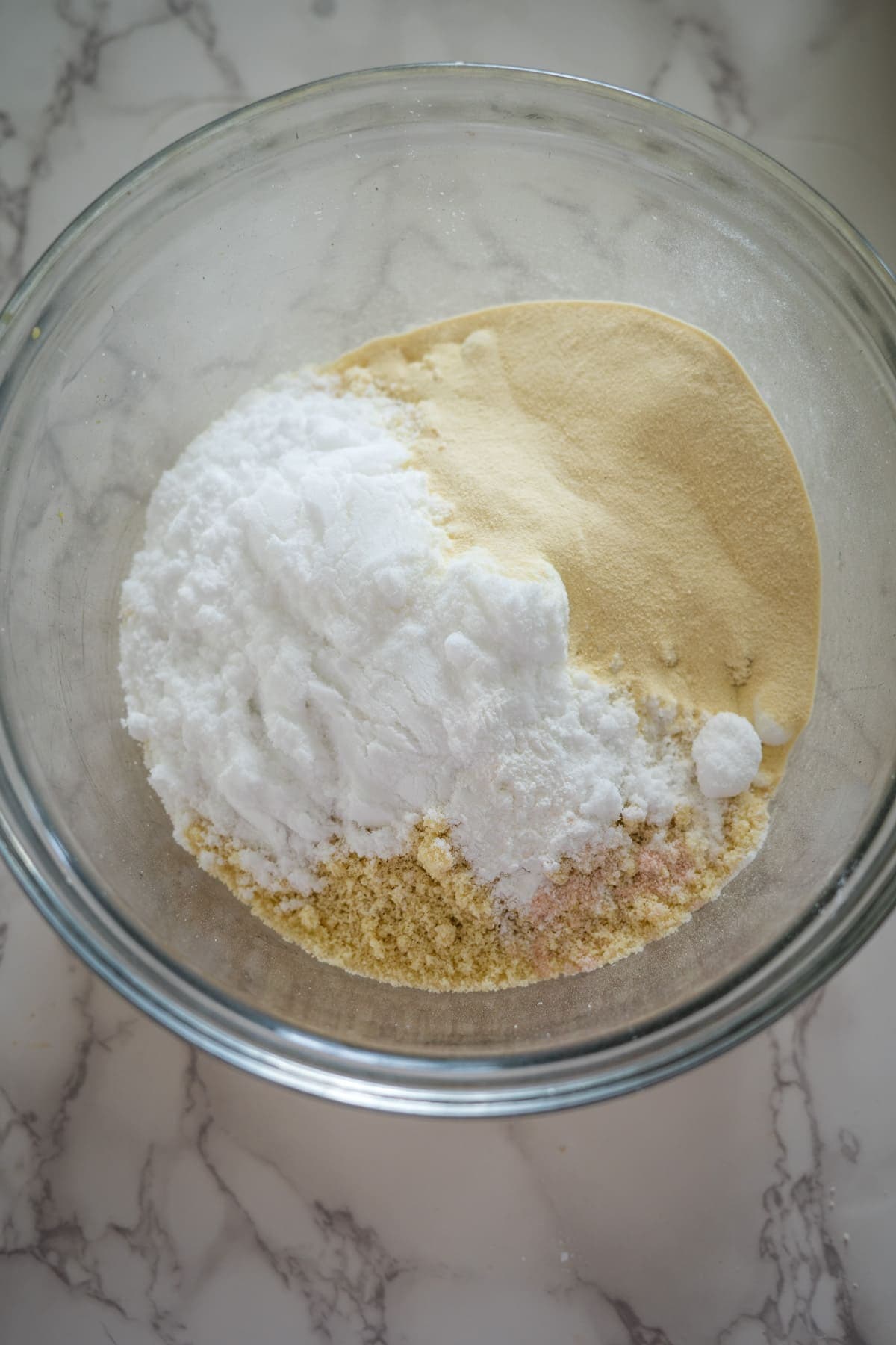 A glass bowl containing a mixture of various dry ingredients, including flour, sugar, and possibly baking powder for cottage cheese cookies, sits on a marble countertop.