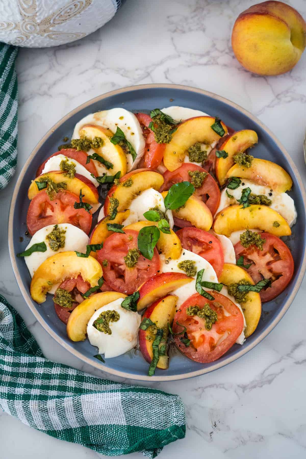 A plate of caprese salad with alternating slices of fresh tomatoes, mozzarella cheese, and peaches, garnished with basil and drizzled with pesto.