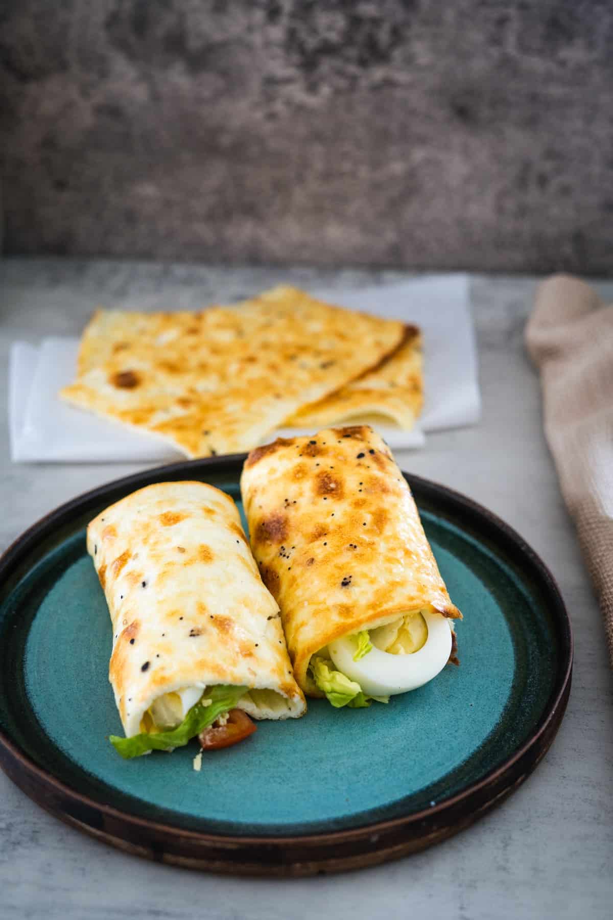 A teal plate with two rolled flatbreads filled with lettuce, egg, and a hint of cottage cheese, placed on a light gray tabletop. In the background, more flatbreads are partially visible on a white napkin.