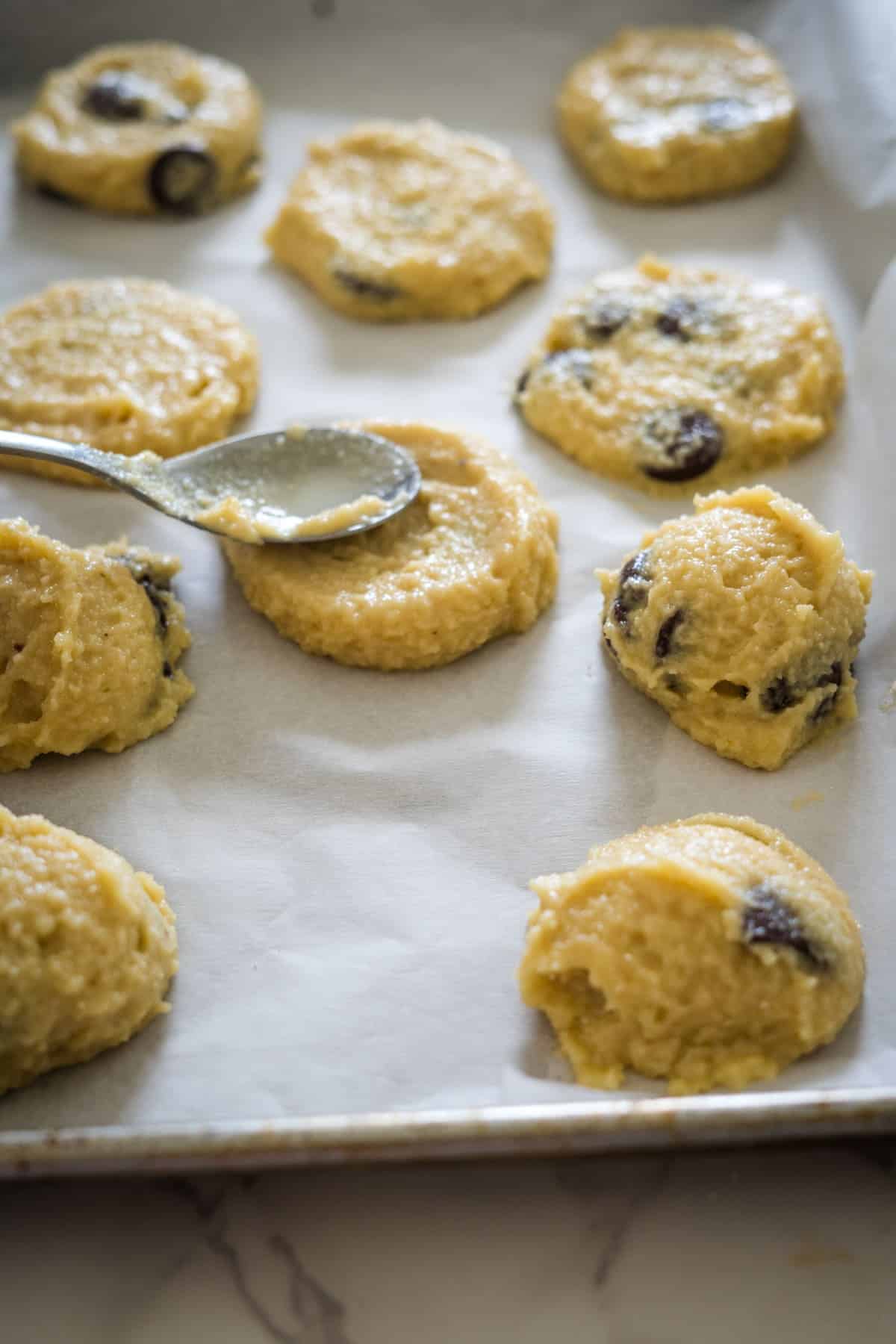 Cookie dough with chocolate chips is placed on a parchment-lined baking tray, resembling the process of making cottage cheese cookies, with a metal spoon used for scooping the dough.