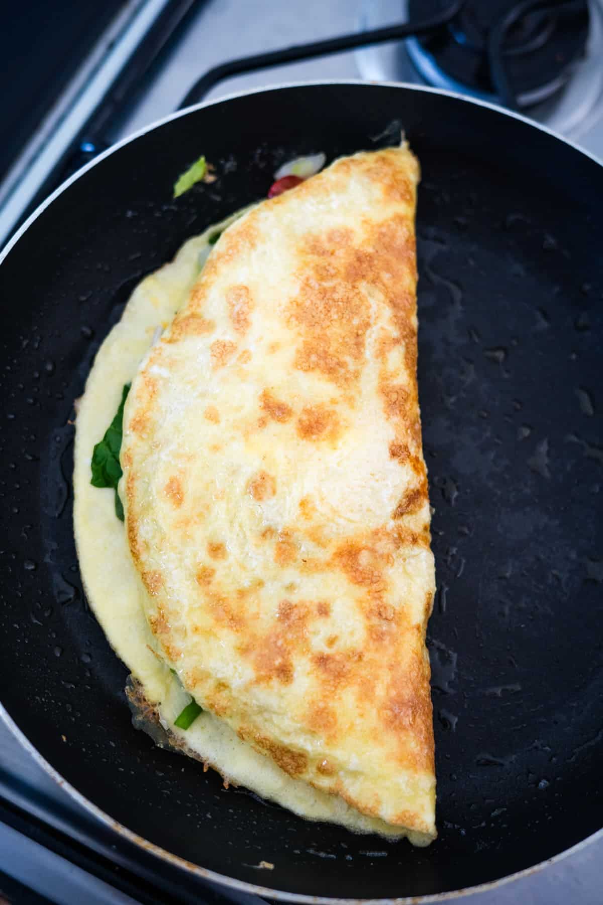 Golden-brown omelette with visible greens, half-folded in a black frying pan on a stove.