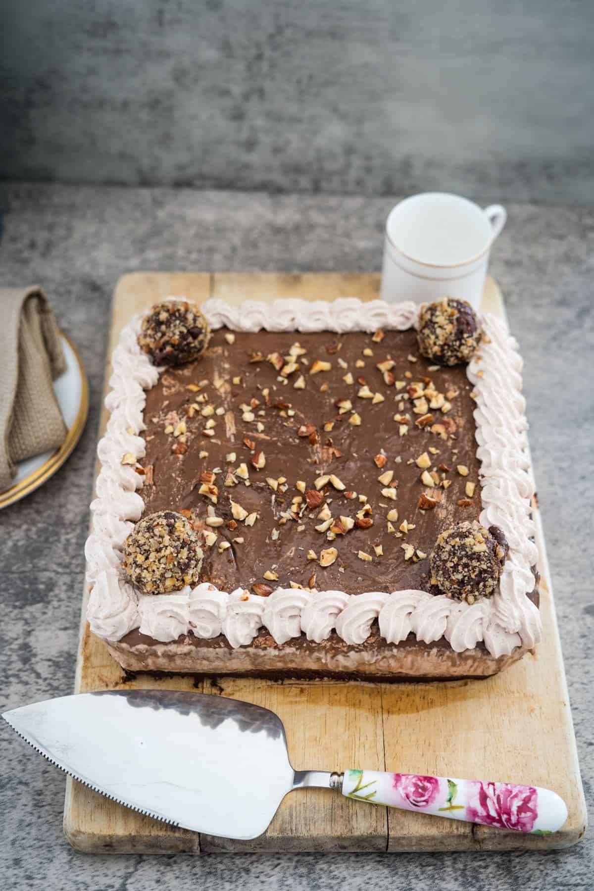 A rectangular chocolate cake with a piped frosting border, chopped nuts, and four round truffles on top. A cake server with a floral handle and a white mug are nearby on a wooden board and gray surface.