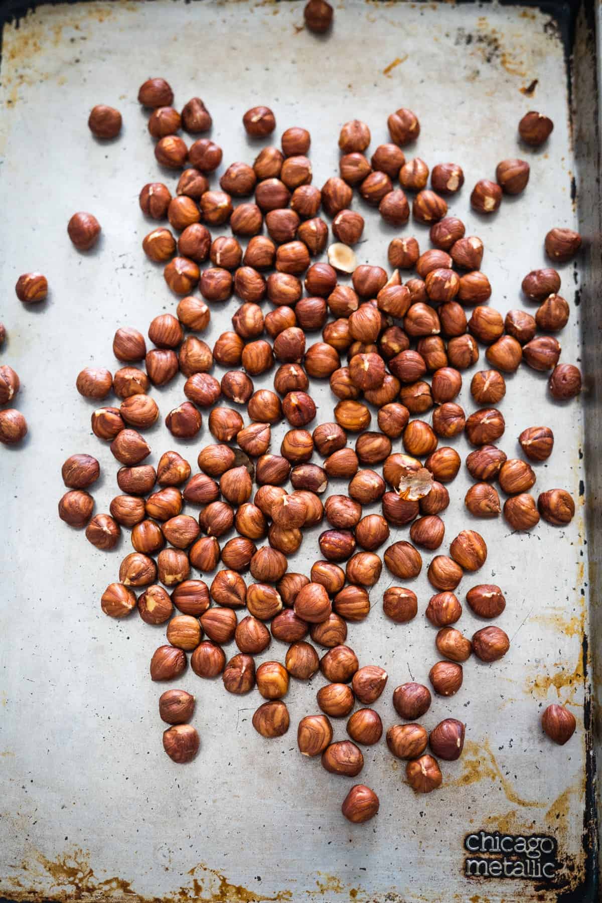 A baking sheet with a spread of raw hazelnuts, some clustered together. The surface is lightly stained, and the bottom right corner has "chicago metallic" branding. Perfect for creating your own sugar-free Nutella at home.