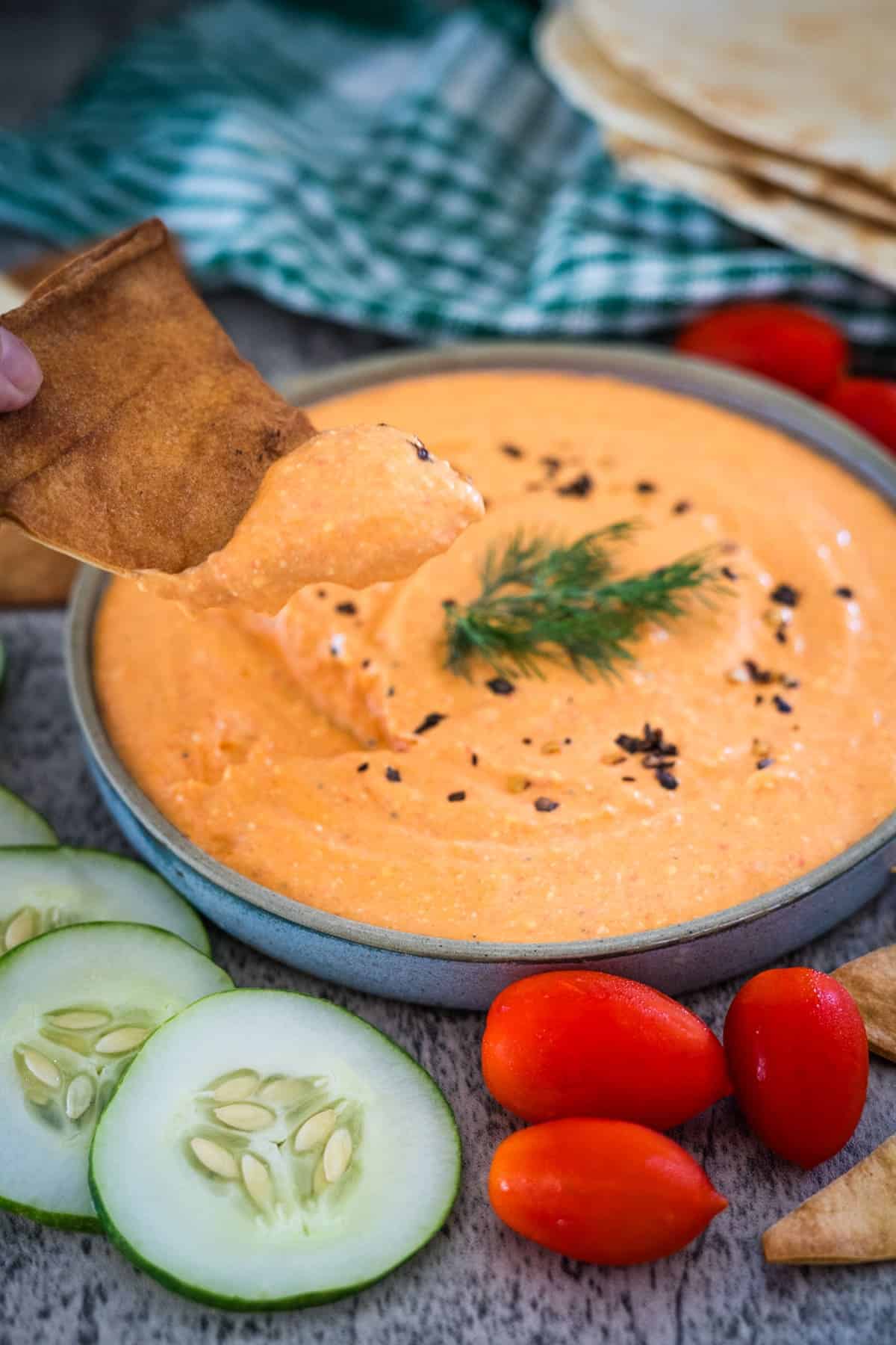 A bowl of creamy feta pepper dip garnished with a sprig of dill, surrounded by cucumber slices, cherry tomatoes, and pita chips. A hand dips a chip into the dip.