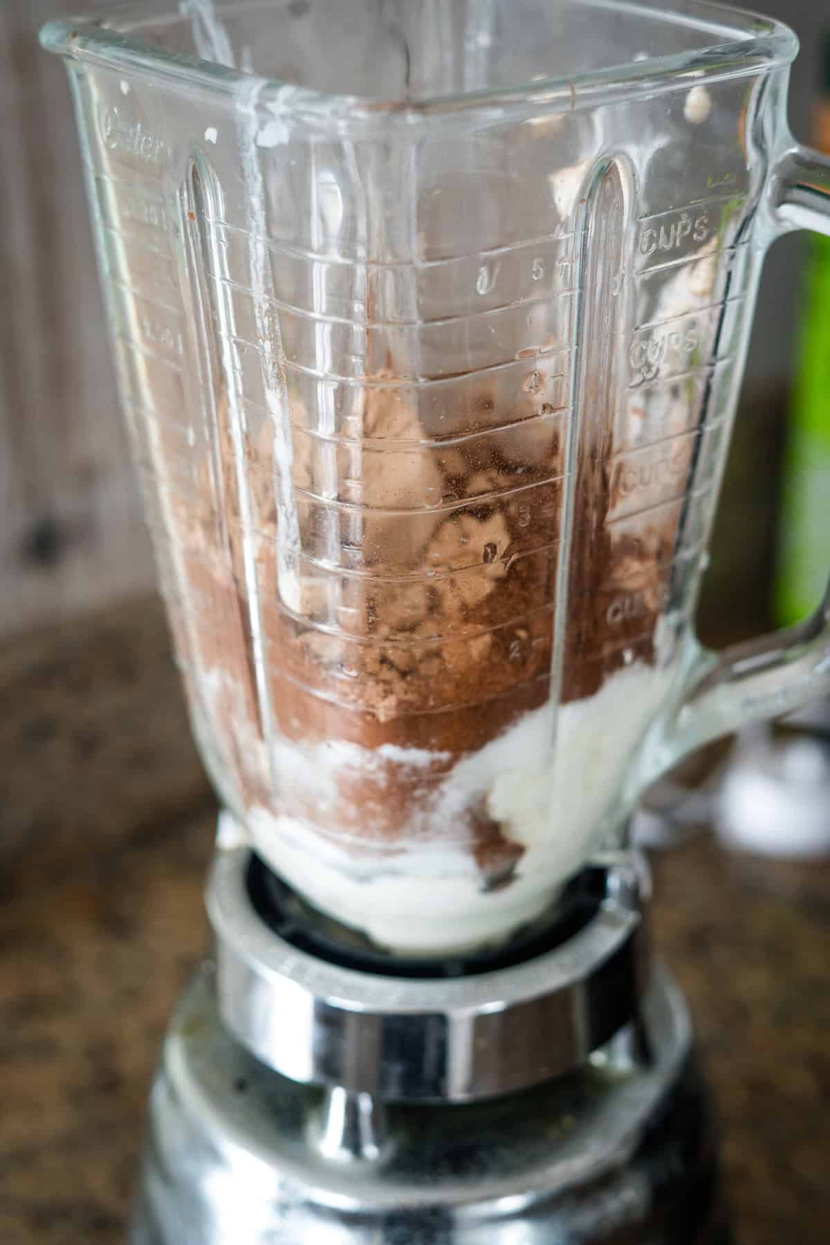 A blender containing cocoa powder, yogurt, and other ingredients is placed on a countertop, ready to blend.