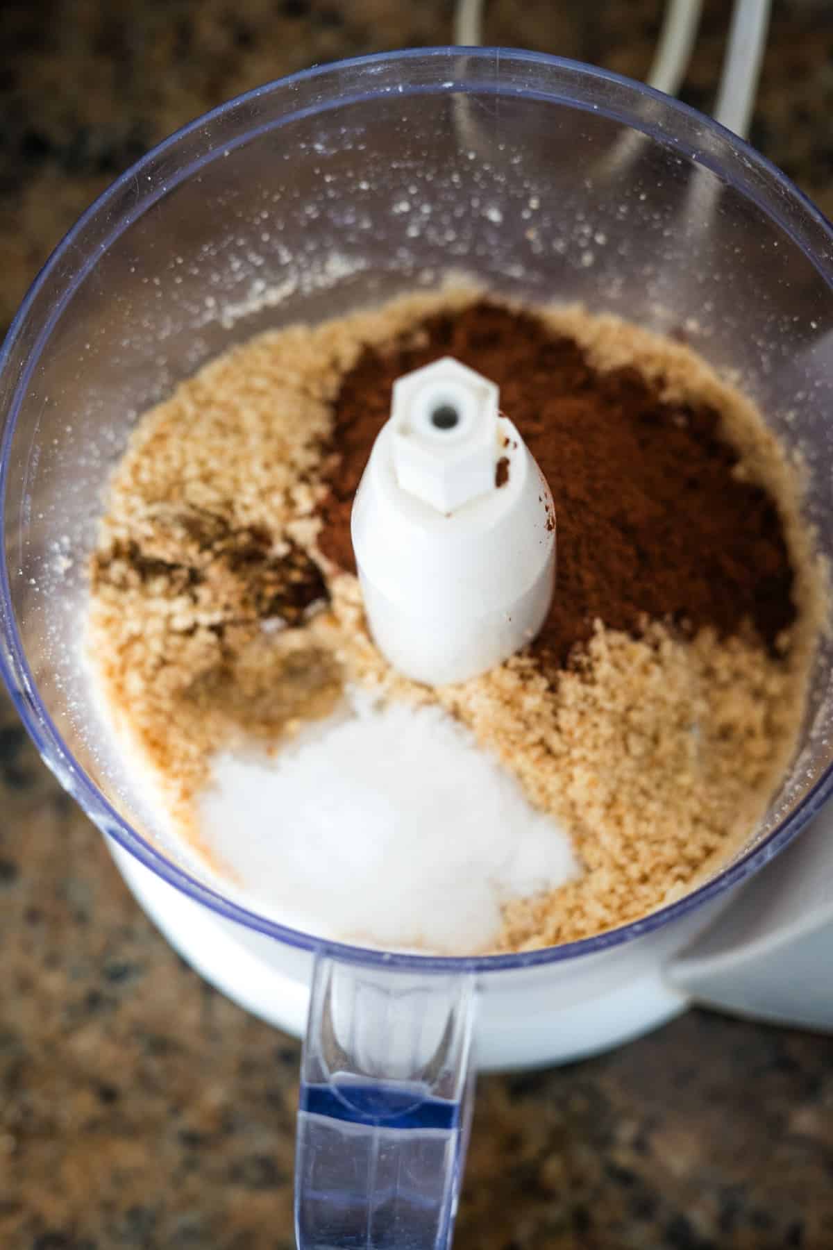 A food processor bowl containing ground almonds, cocoa powder, coconut, and other ingredients is ready for blending on a kitchen countertop, promising a delightful batch of sugar-free Nutella.