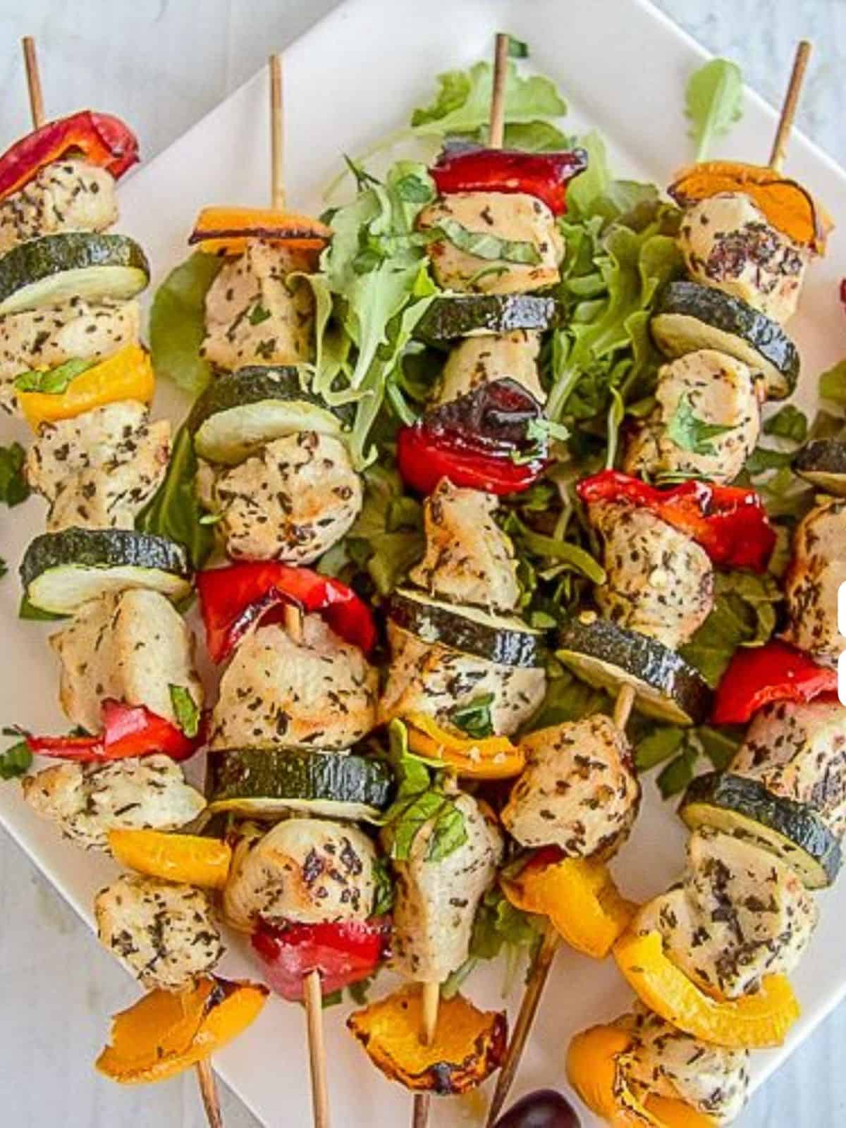 Skewers of grilled chicken kofta kebabs and vegetables, including bell peppers and zucchini, arranged on a bed of leafy greens on a white plate.