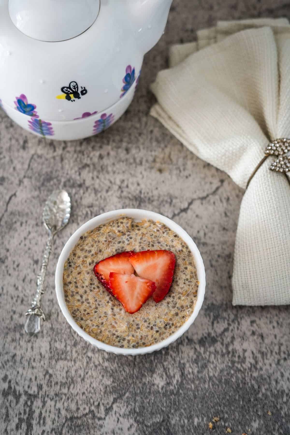 A bowl of chia and flaxseed pudding topped with sliced strawberries on a textured gray surface, near a white teapot and a napkin.