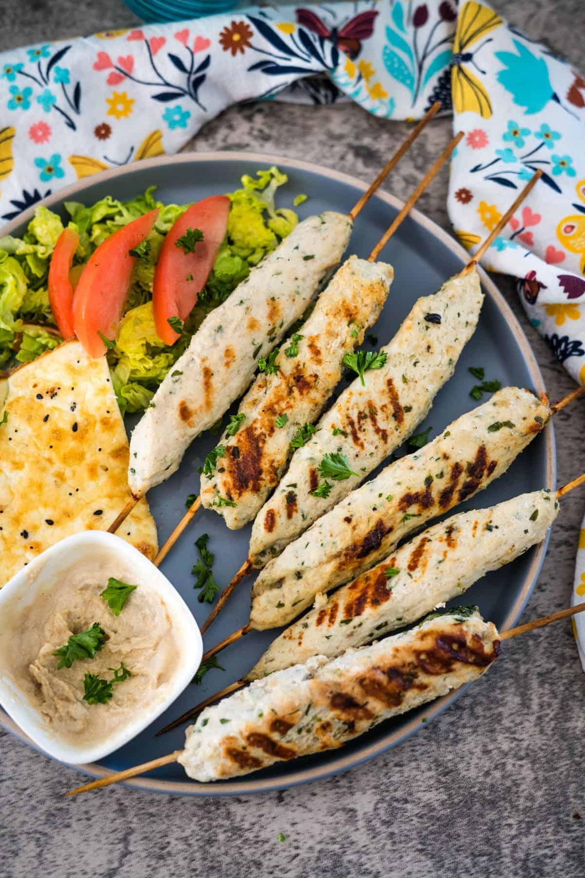 A plate of chicken kofta kebabs with a side of hummus, lettuce, tomato slices, and flatbread. A colorful floral napkin is in the background.