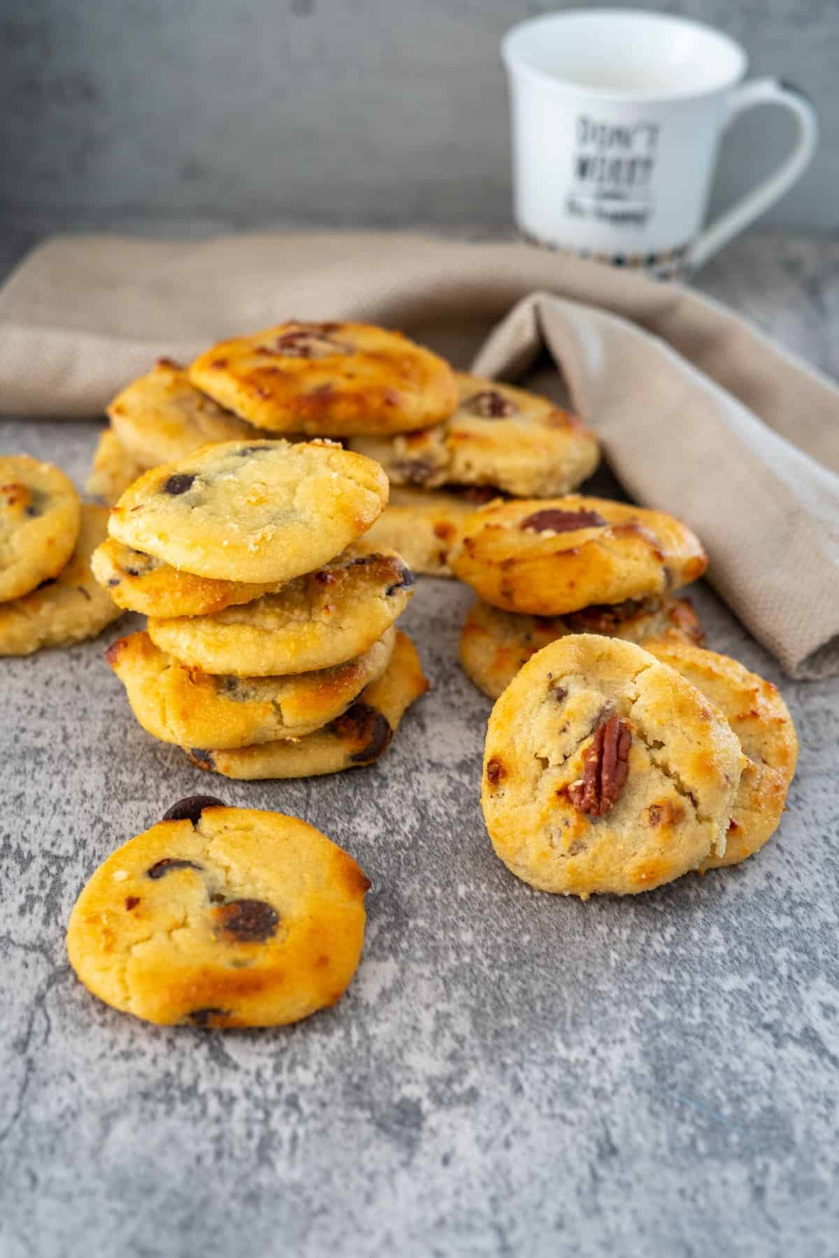 A stack of cottage cheese cookies with chocolate chips and nuts is placed in front of a white mug and a beige napkin on a grey textured surface.