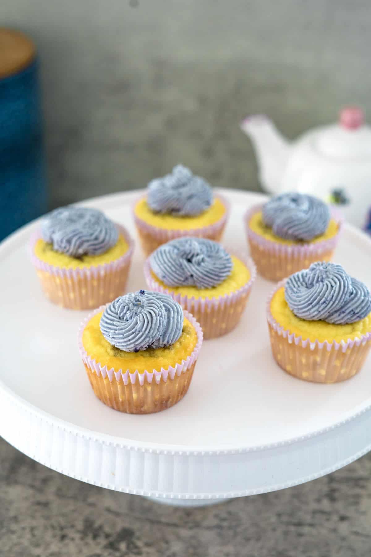 A white cake stand holds six cupcakes with yellow bases and blue-gray frosting swirls. A teapot is partially visible in the background.