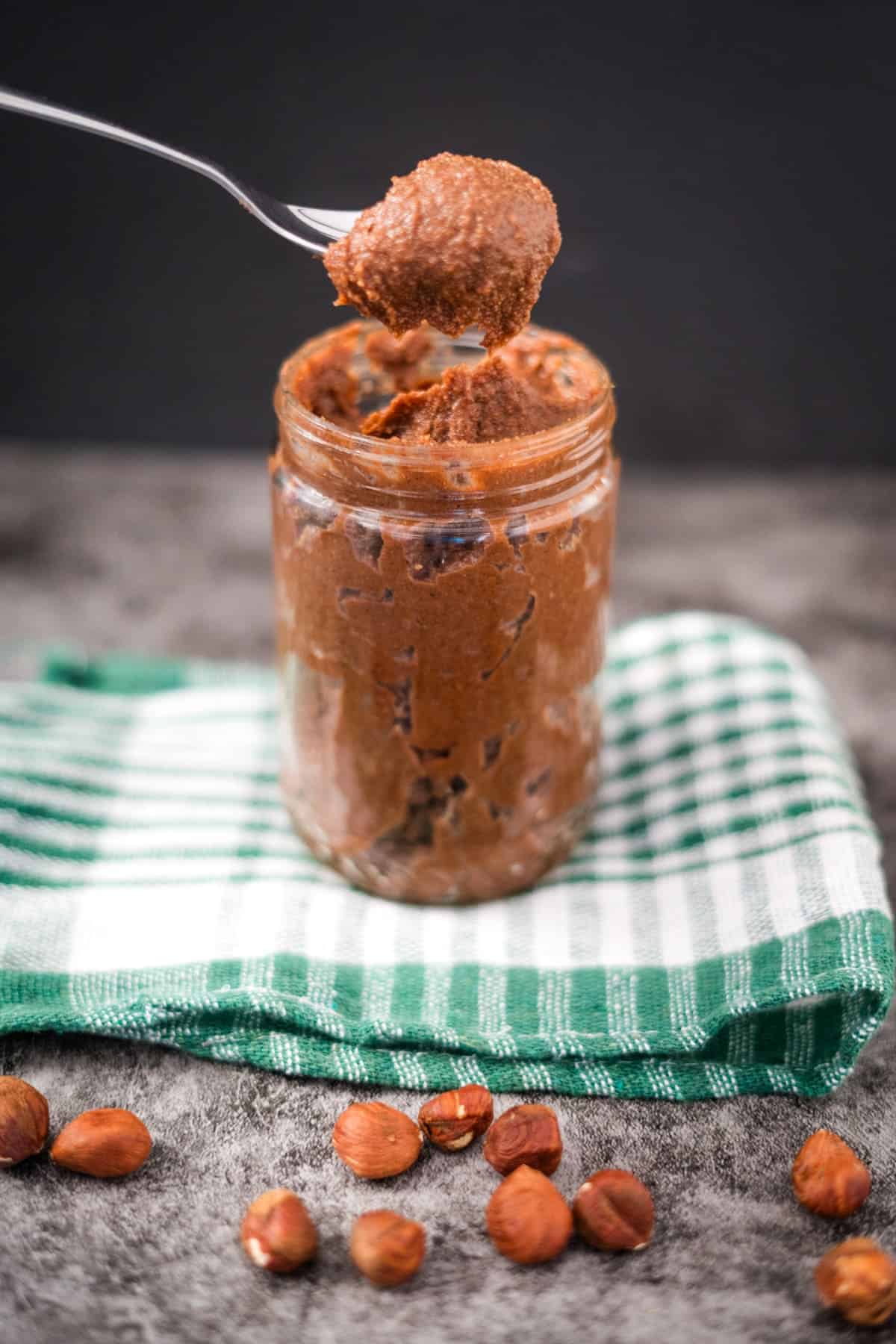 A jar of sugar-free Nutella with a portion scooped on a spoon above the jar, set on a green and white checkered cloth with hazelnuts scattered around.