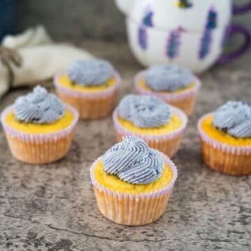 Six yellow cupcakes with light blue swirled frosting are arranged on a textured surface with a decorated teapot in the background.