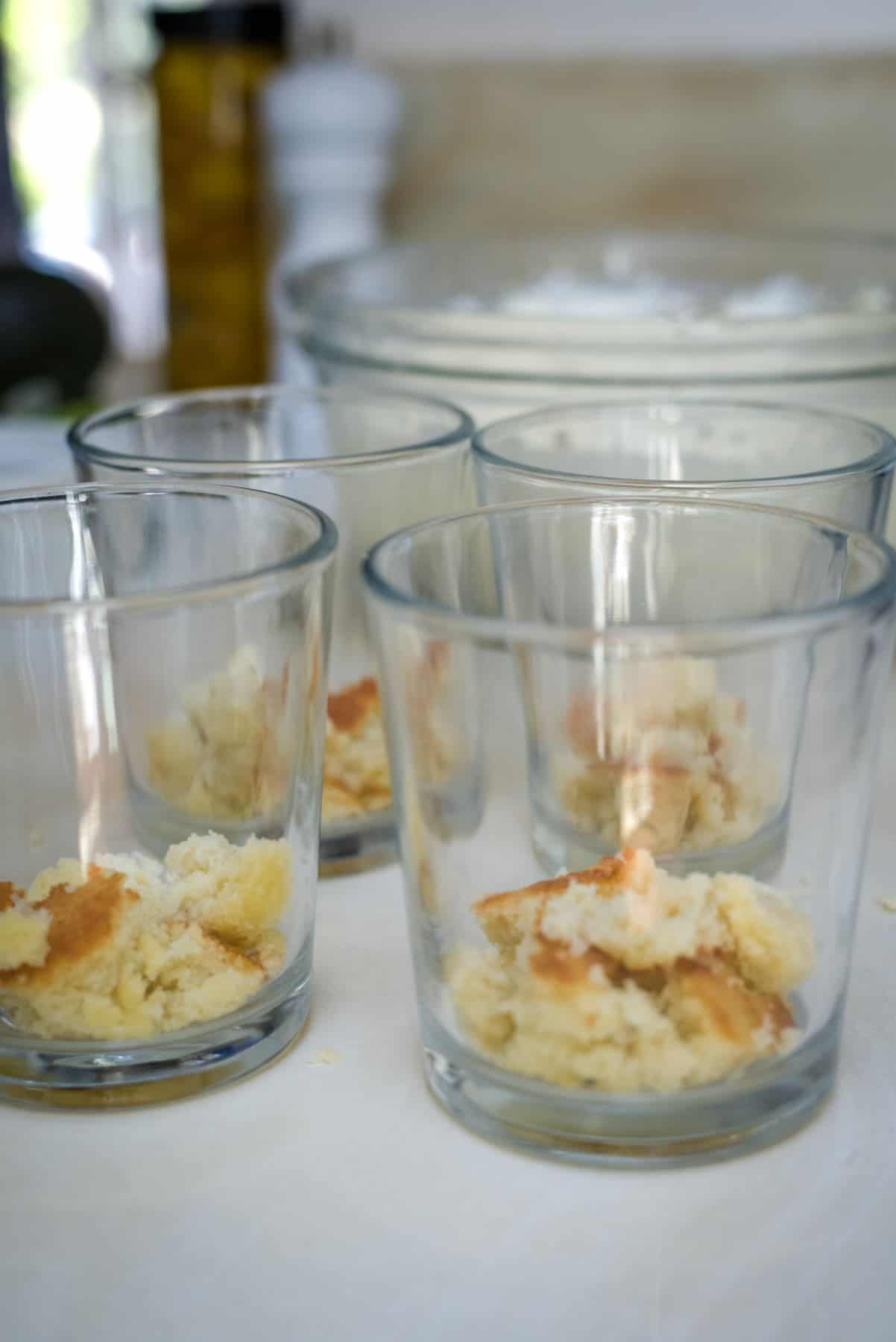 Four clear glasses each contain pieces of crumbled cake. A bowl of flour is visible in the background.