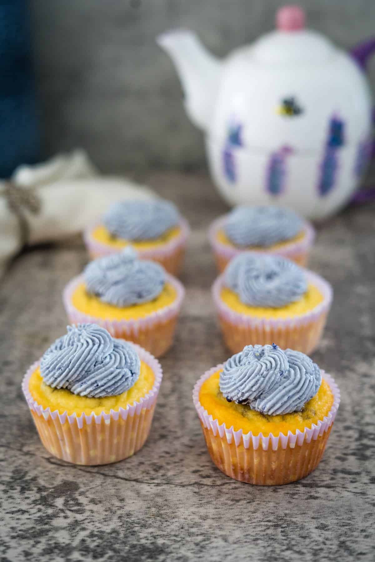 Seven yellow cupcakes with light purple frosting are arranged on a countertop with a white teapot in the background.