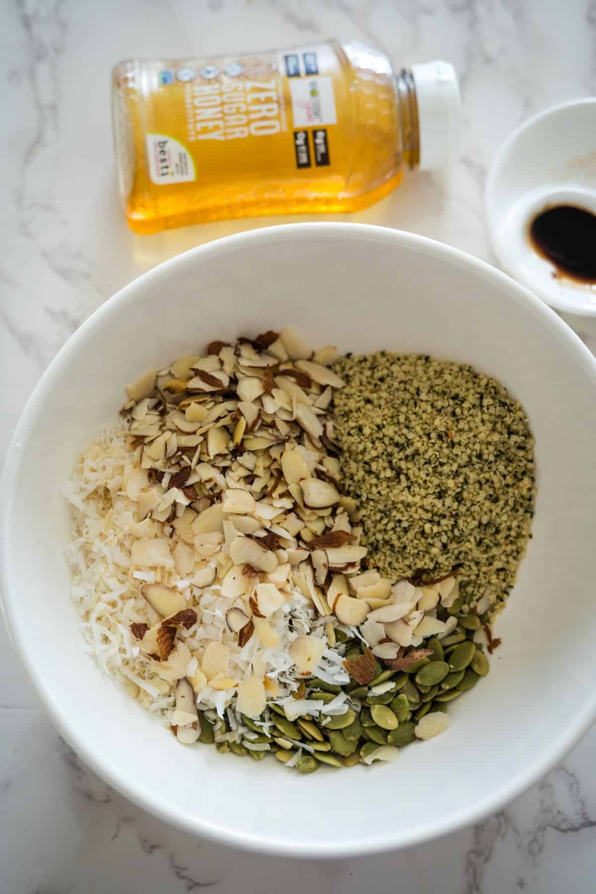 A bowl containing a mix of sliced almonds, shredded coconut, and pumpkin seeds—ingredients for keto breakfast cookies—with a bottle of honey and a small dish of soy sauce in the background.