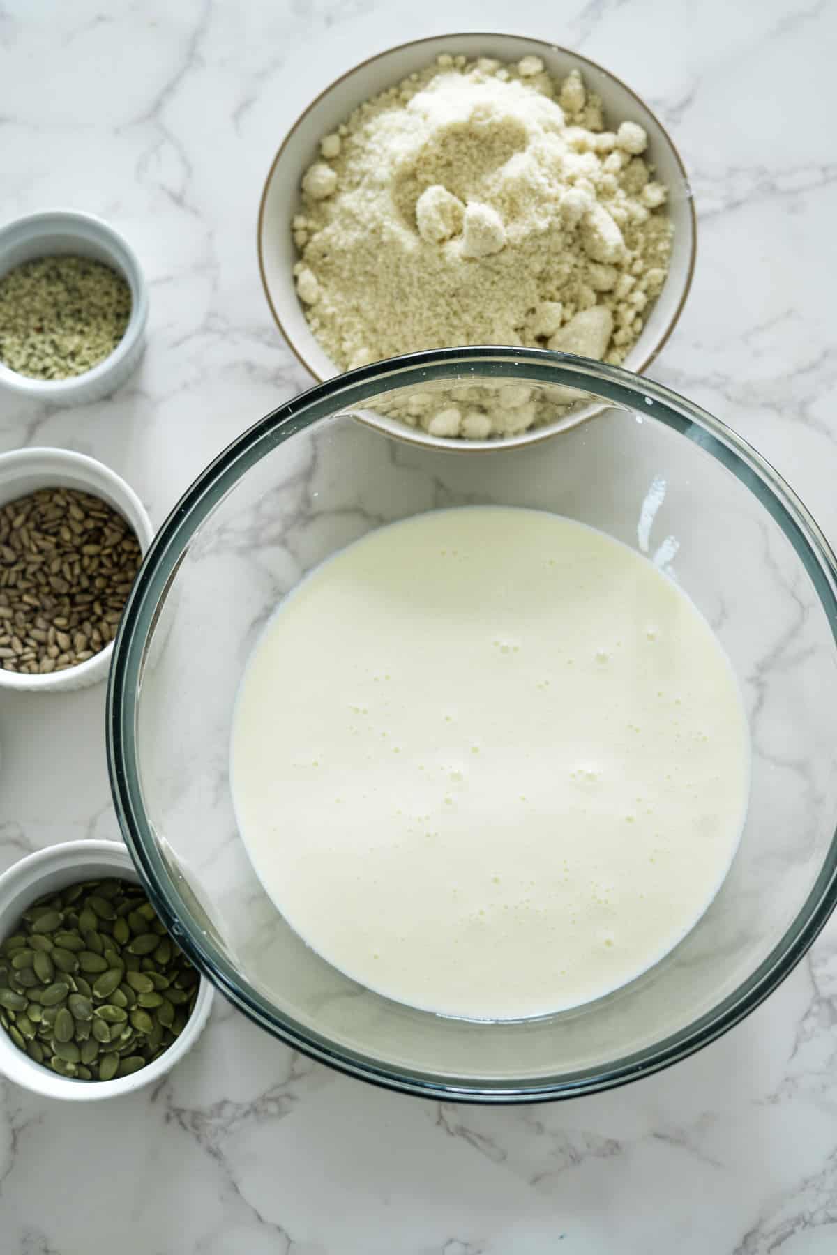 A glass bowl filled with a creamy liquid is placed on a marble surface, surrounded by four smaller bowls containing various seeds and a larger bowl with powdery ingredients.