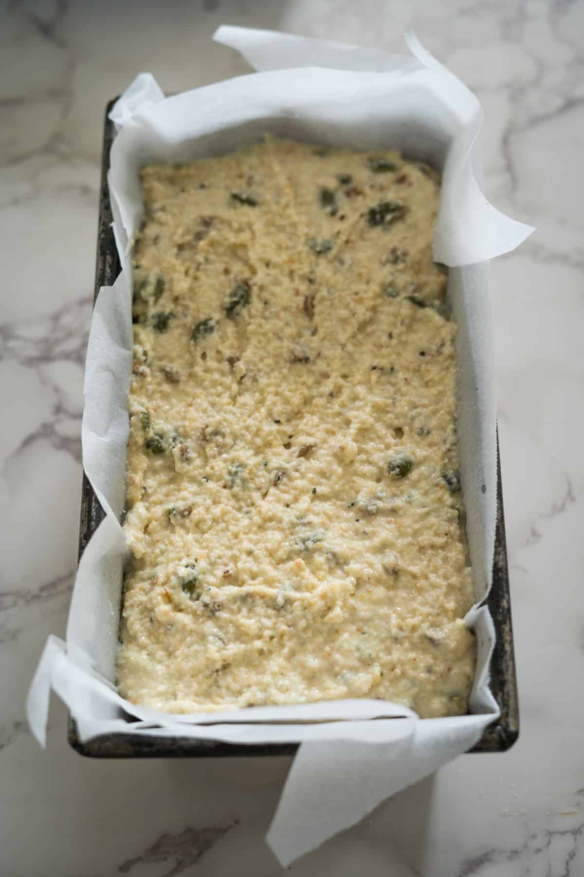 A loaf pan filled with uncooked cornbread batter lined with parchment paper rests on a marble surface.