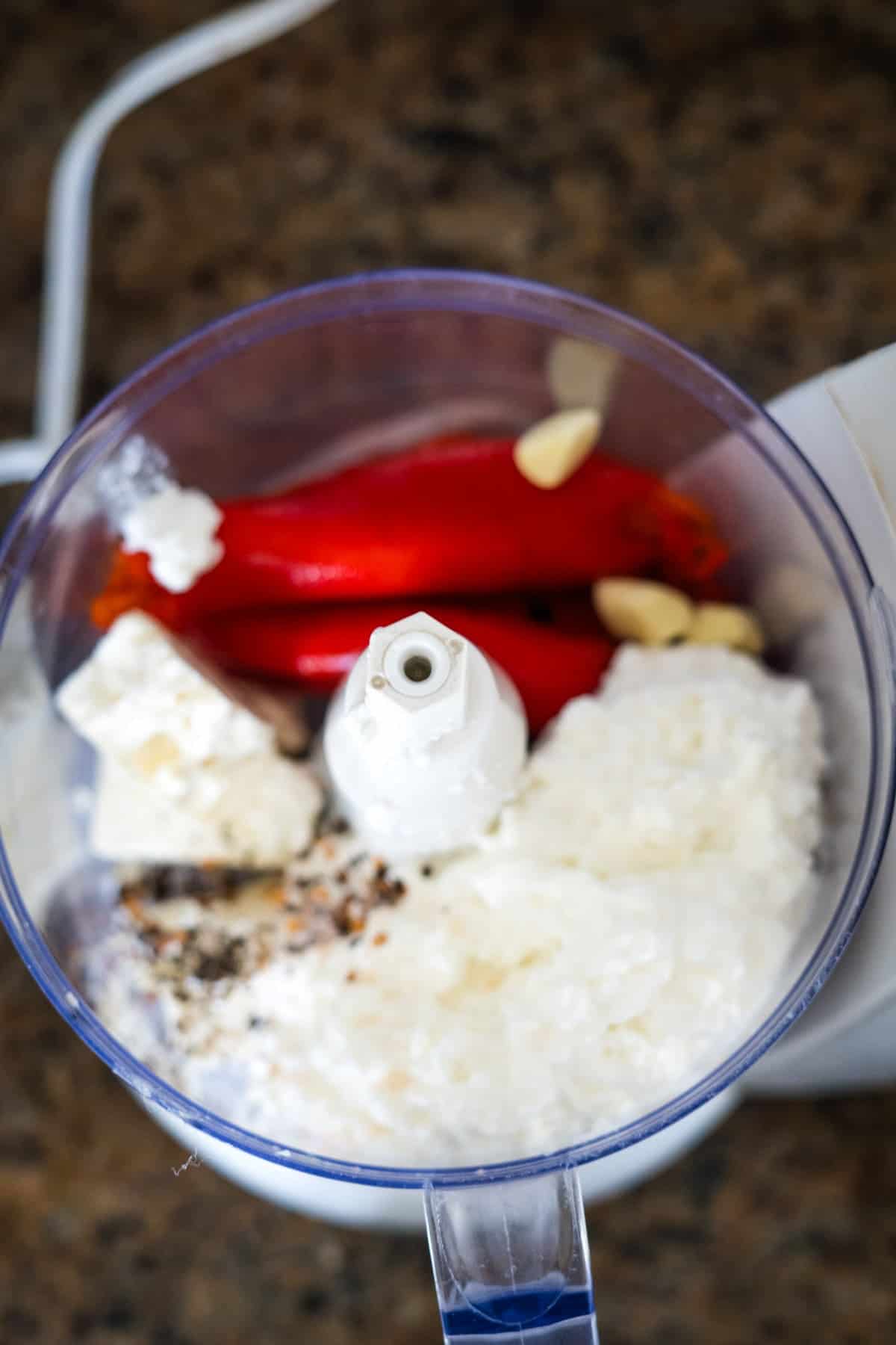 A top view of a food processor containing red bell peppers, garlic cloves, feta cheese, pepper, and other ingredients, set on a granite countertop.