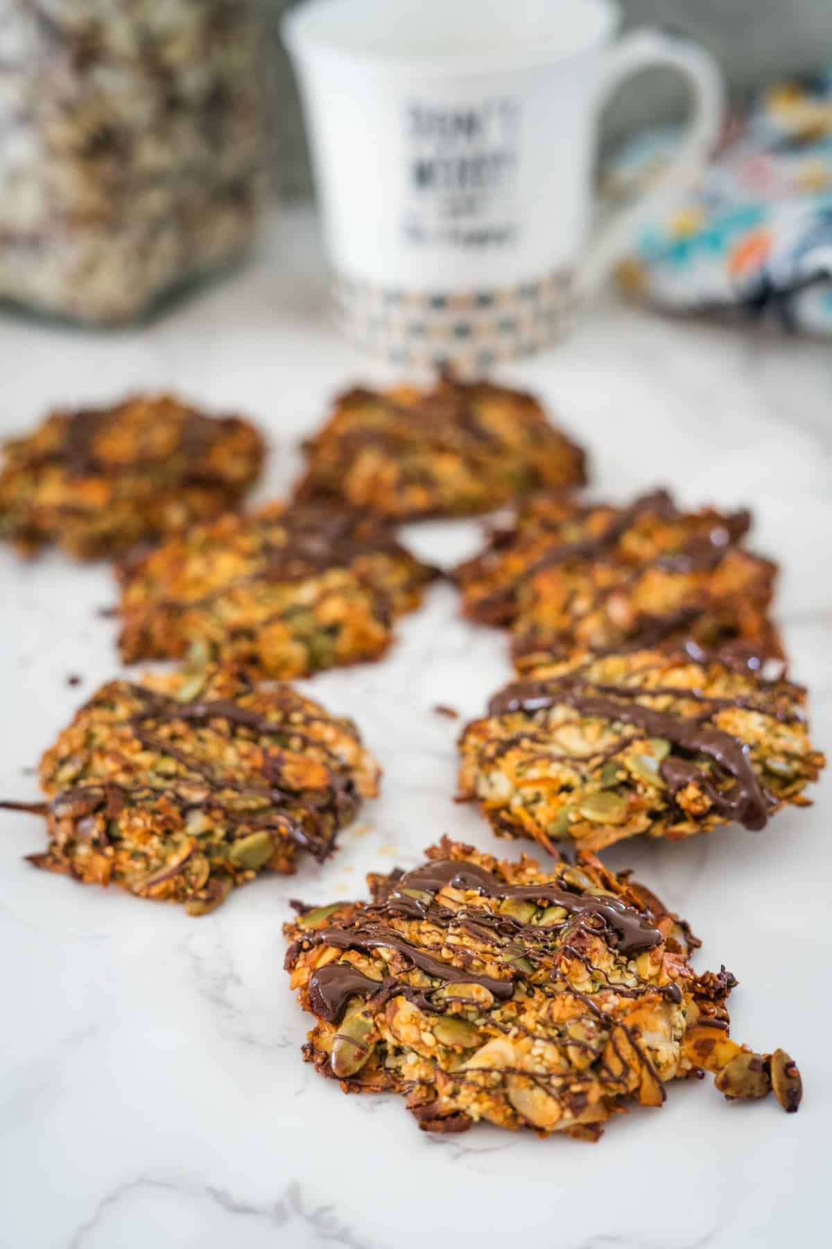 Homemade granola cookies drizzled with chocolate on a marble countertop, with a mug and dried flowers in the background.