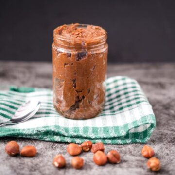 A jar of hazelnut spread is placed on a green and white checkered cloth, with a spoon and scattered hazelnuts in front.