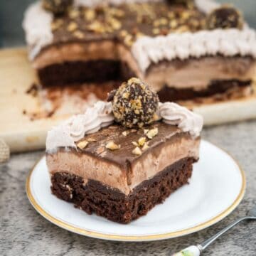 A slice of chocolate and cream layered dessert with a nut topping on a white plate, with the larger portion in the background on a wooden board.