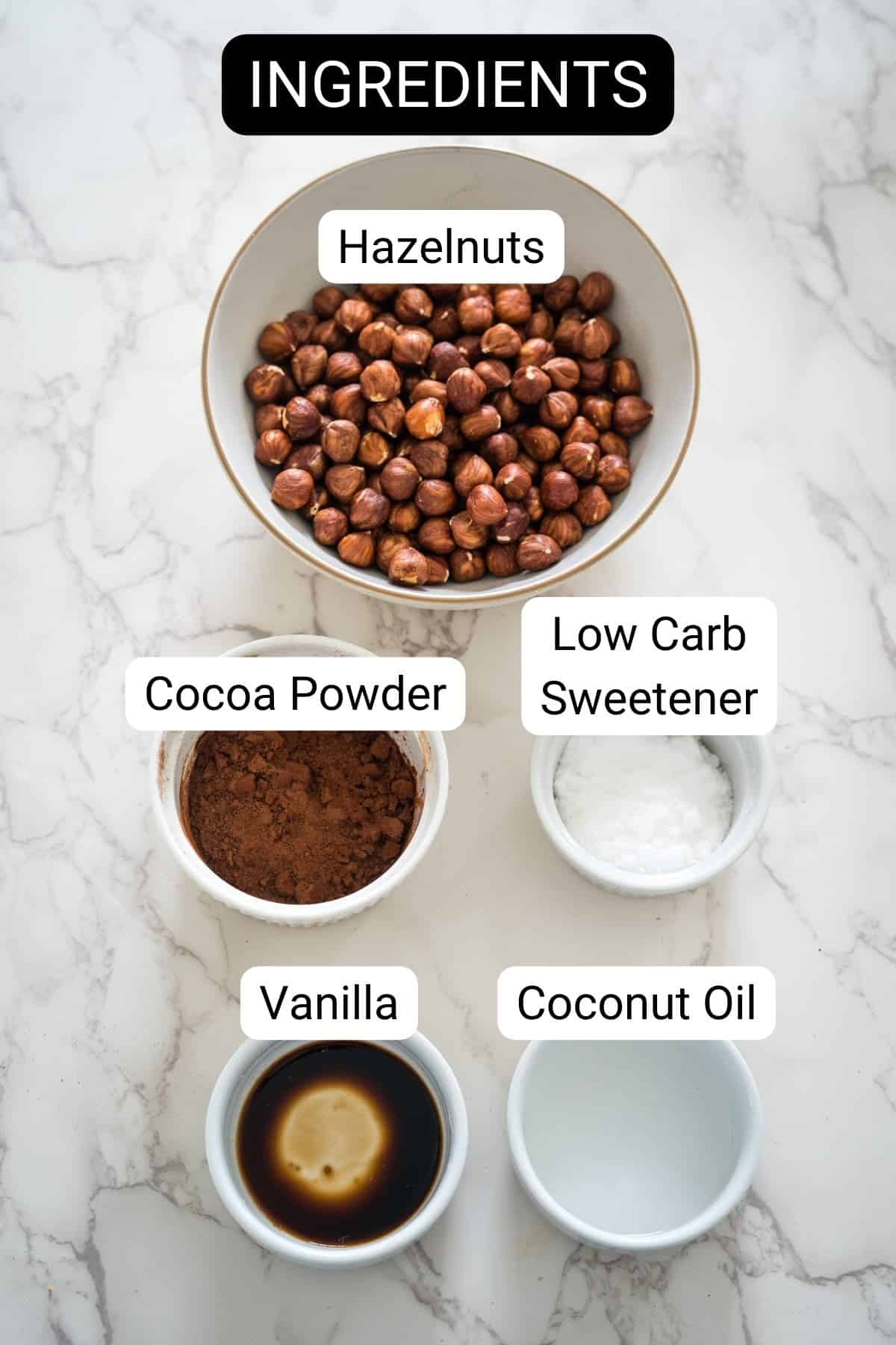 Top view of ingredients in bowls labeled: hazelnuts, low carb sweetener, cocoa powder, coconut oil, and vanilla. Text "INGREDIENTS" at the top. All placed on a marble surface, perfect for making sugar free nutella.