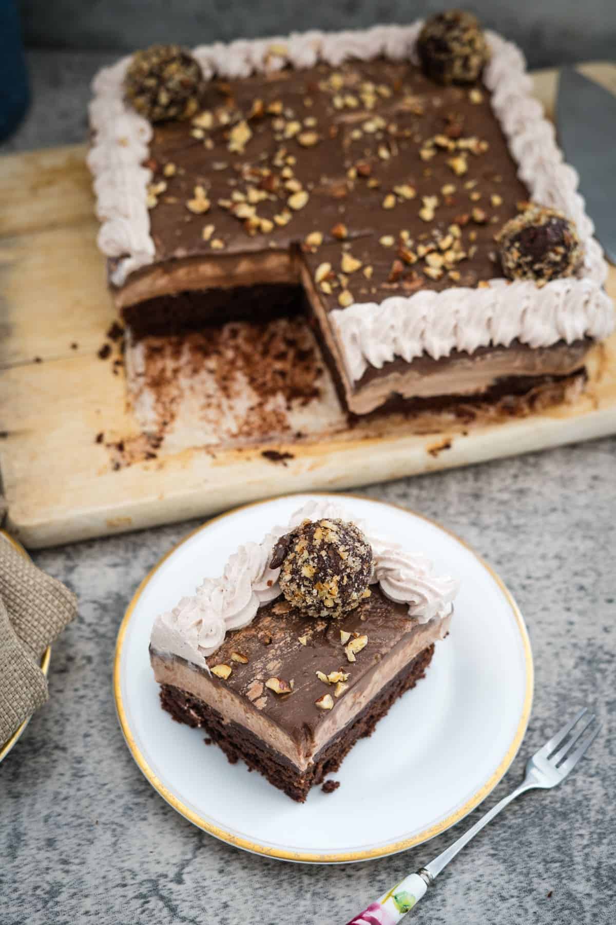 A slice of chocolate cake with cream topping and a chocolate truffle on top is placed on a plate. The remaining Nutella brownie cheesecake with a similar topping is on a wooden board in the background.