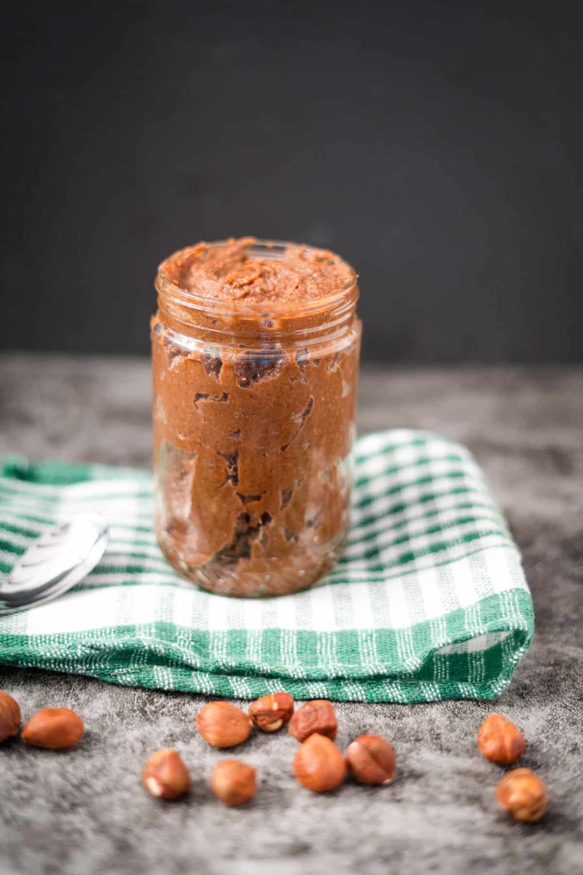 A jar filled with sugar-free Nutella sits on a green and white checkered cloth, with a spoon and scattered hazelnuts around it.