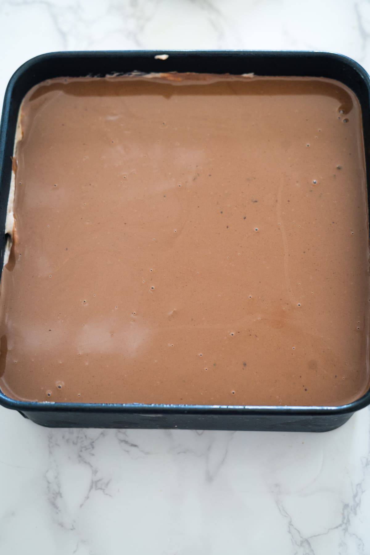 A black baking pan is filled with a smooth layer of Nutella brownie cheesecake batter, resting on a white marble surface.