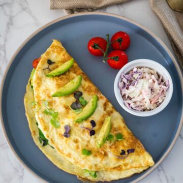 A folded omelette garnished with avocado slices and herbs on a blue plate, accompanied by a side of coleslaw and cherry tomatoes.