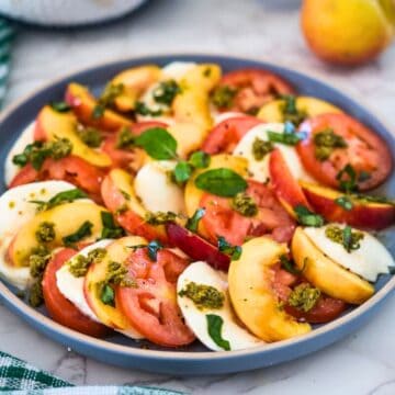 A colorful caprese salad with sliced tomatoes, peaches, and mozzarella, garnished with basil and drizzled with pesto.