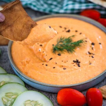 A hand dipping a piece of pita into a bowl of creamy orange dip garnished with a sprig of dill. Sliced cucumbers and cherry tomatoes are visible around the bowl.