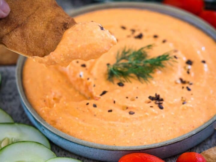 A hand dipping a piece of pita into a bowl of creamy orange dip garnished with a sprig of dill. Sliced cucumbers and cherry tomatoes are visible around the bowl.