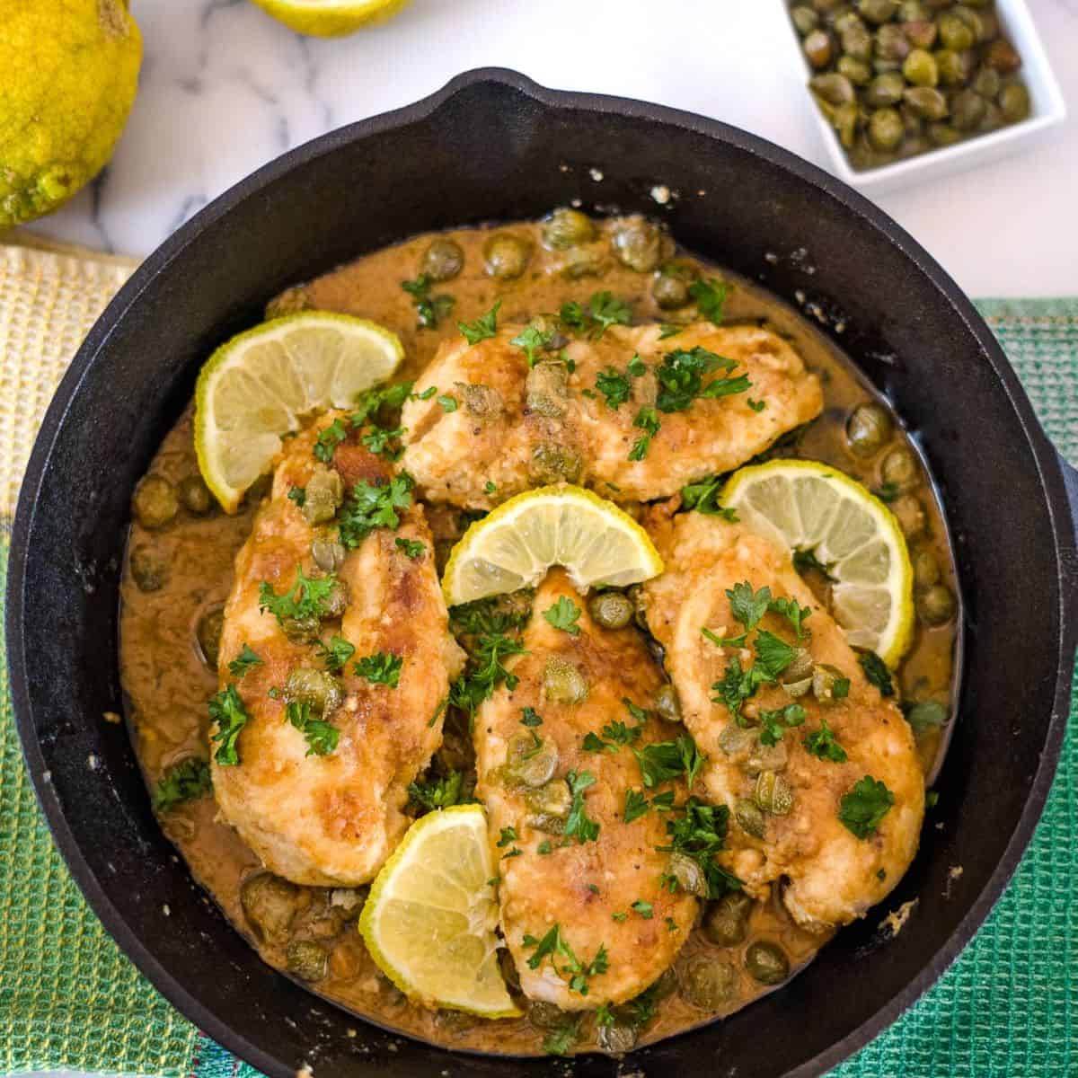 A skillet containing four cooked chicken breasts garnished with lemon slices, capers, and chopped parsley, in a brownish sauce. A white dish with capers and half a lemon are seen nearby.