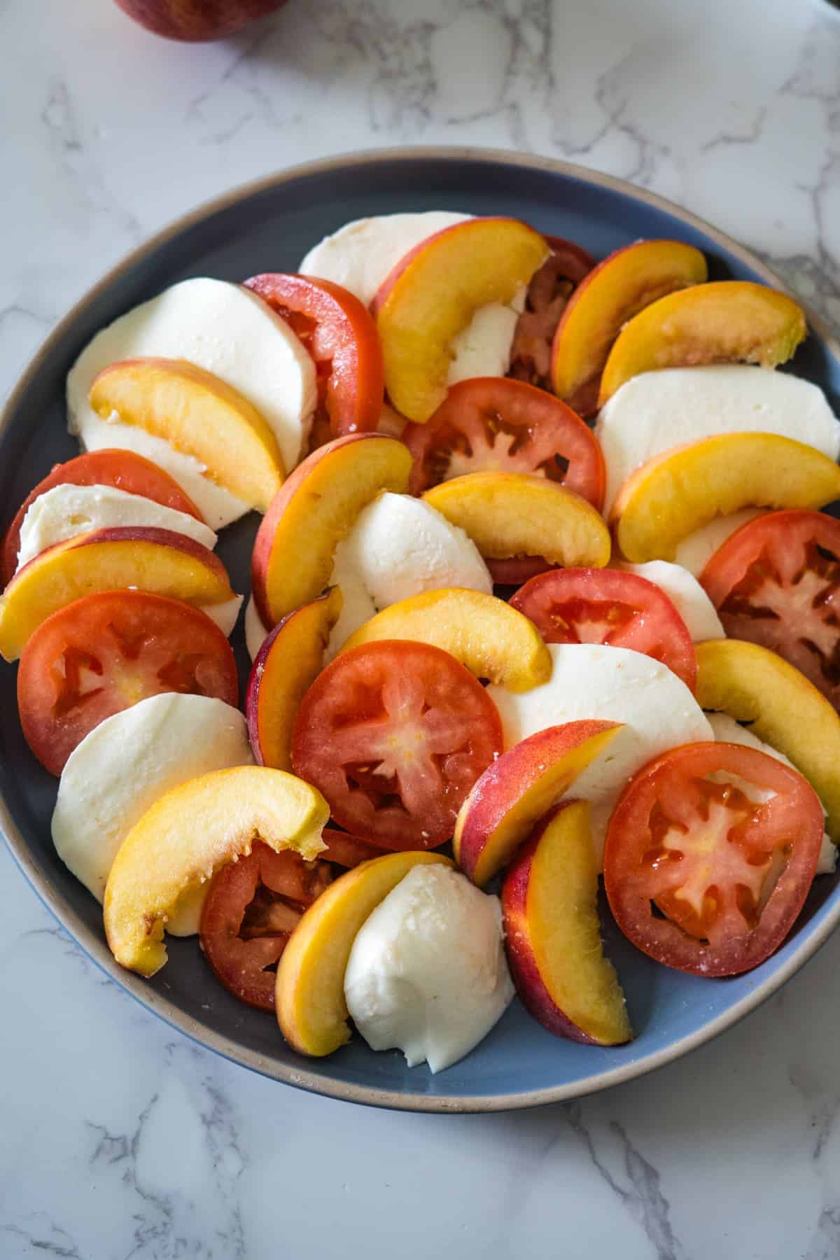 A plate featuring slices of tomato, mozzarella cheese, and peaches arranged alternately on a marble countertop.