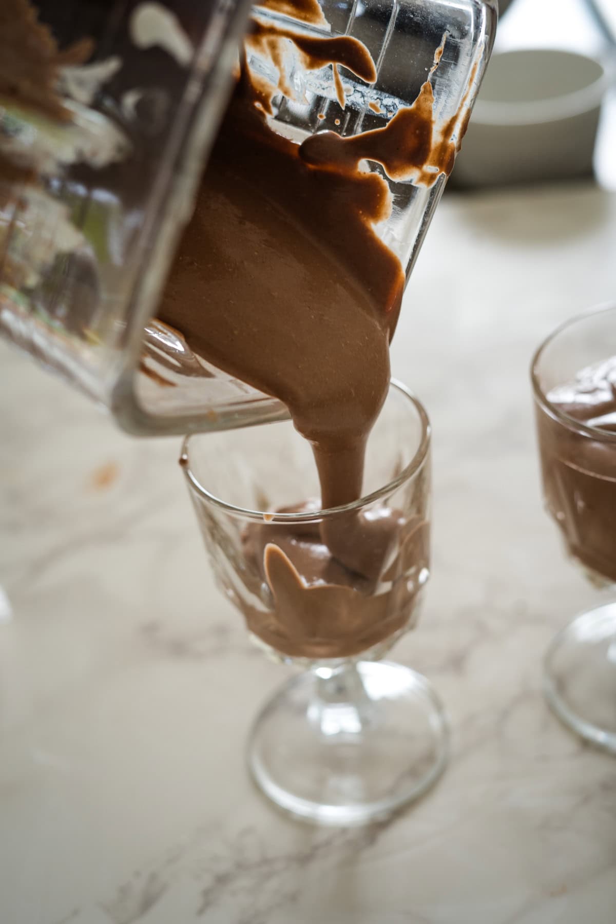 A thick chocolate smoothie is being poured from a blender into a glass cup on a marble countertop. Another glass with the smoothie is visible in the background.